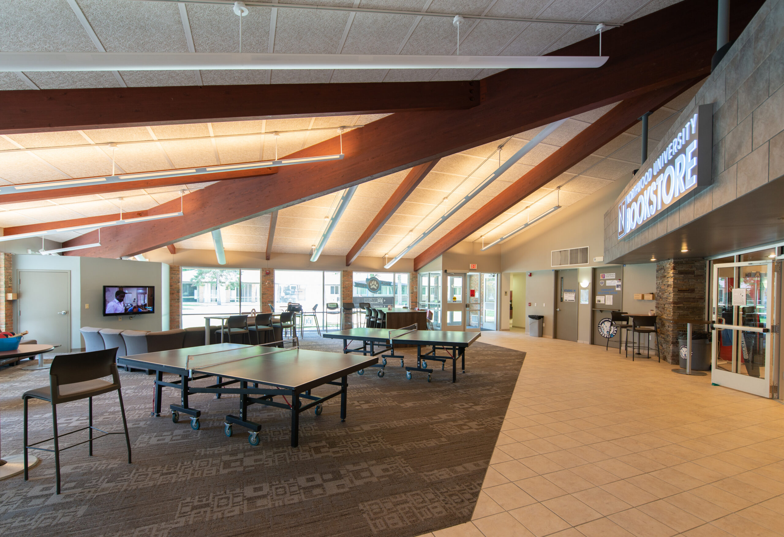 A spacious student center with ping pong tables, chairs, large windows, and a campus bookstore under a sloped ceiling with exposed beams—part of Northwood's Project 100 initiative.