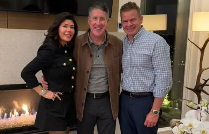 Three people stand side by side indoors near a fireplace, smiling at the camera. A plant and a window with city lights are visible in the background.