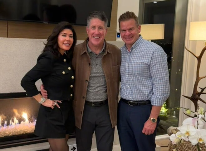 Three people stand side by side indoors near a fireplace, smiling at the camera. A plant and a window with city lights are visible in the background.