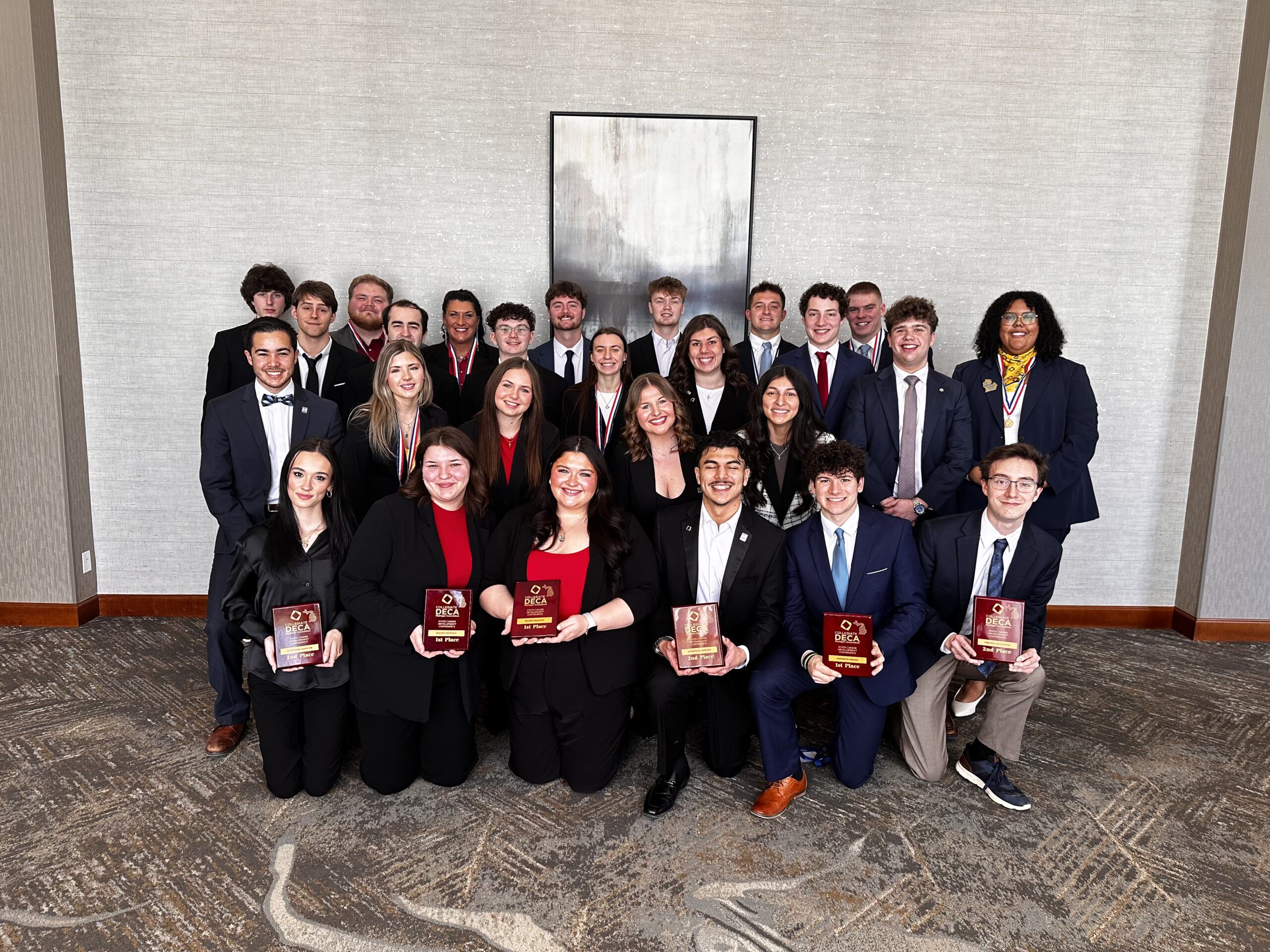 A group of 25 people in business attire pose indoors, with several holding red and gold DECA award plaques, in front of a gray wall and abstract painting.
