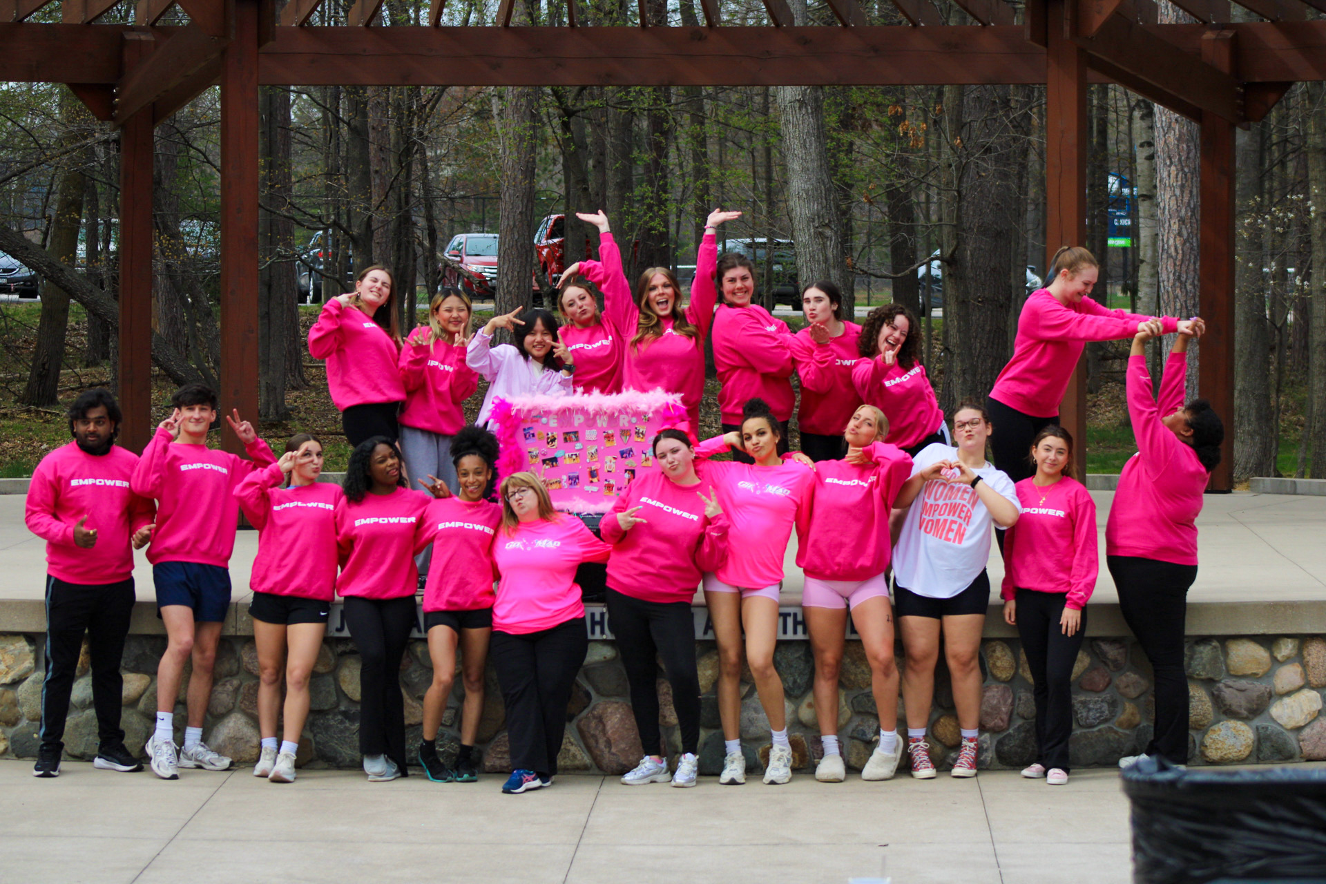 A group of 19 people, mostly women, wear matching pink sweatshirts and pose together outdoors under a wooden pavilion in front of a forested area.