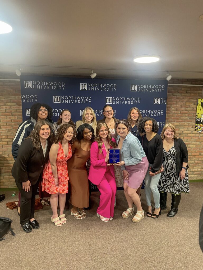 A group of people pose together indoors in front of a Northwood University backdrop, with one person holding a blue plaque or award.