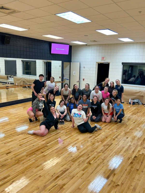 A group of people pose and smile together in a dance studio with mirrors, wooden floors, and a screen displaying text in the background.