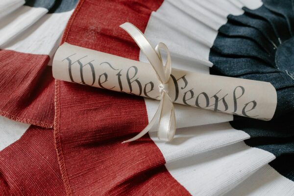 A rolled parchment labeled "We the People," symbolizing freedom, is tied with a ribbon and rests on red, white, and blue fabric resembling the American flag.