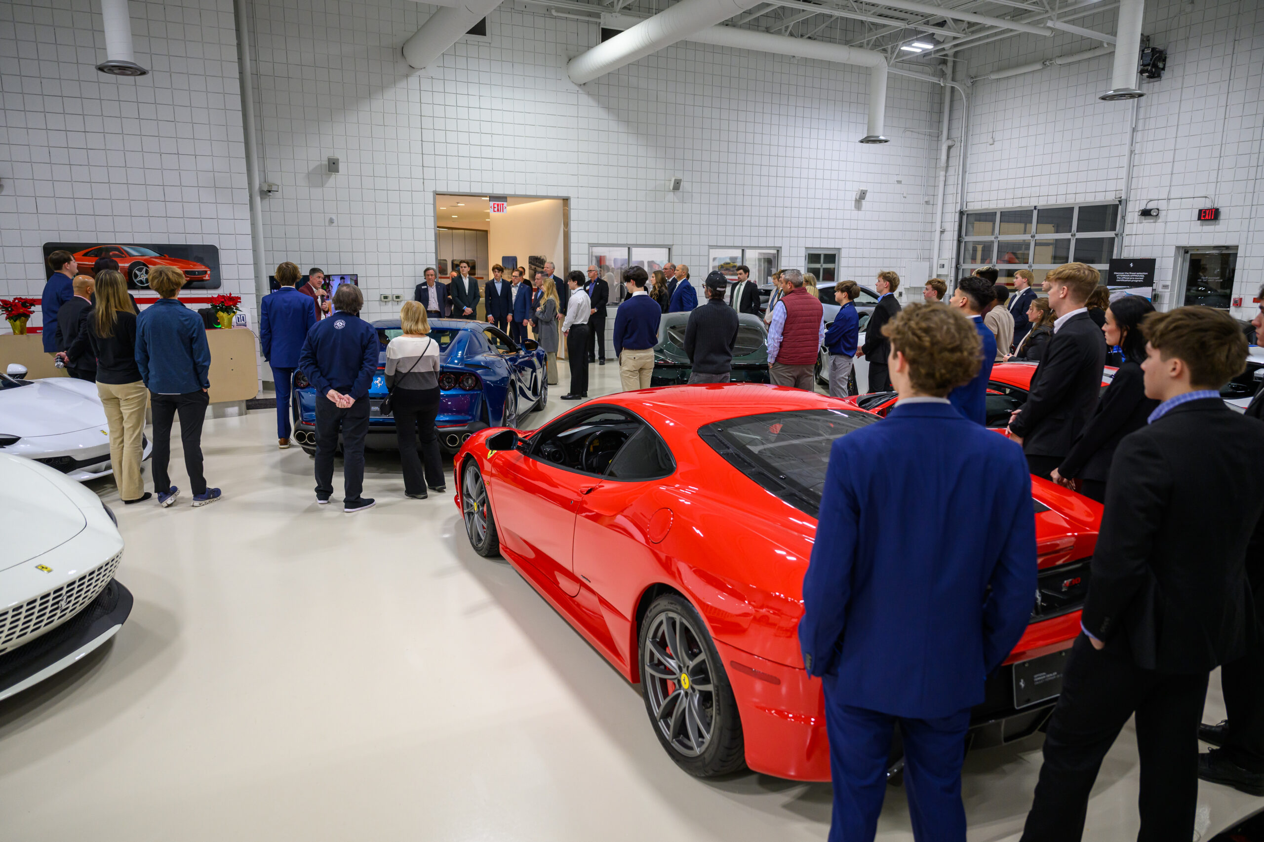A group of people in business attire gather around luxury sports cars inside a brightly lit showroom with white walls and tile flooring, showcasing a collaboration with the university automotive department.