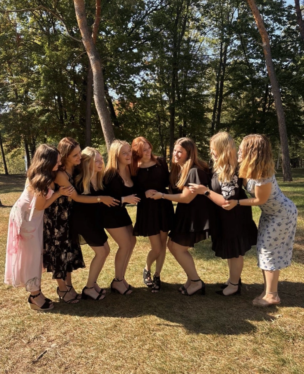 Eight women in dresses stand in a semi-circle outdoors on grass, smiling and holding hands, with trees and sunlight in the background, celebrating their sisterhood in Alpha Gamma Delta.