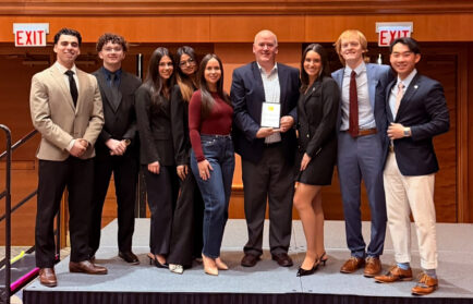 Nine people, formally dressed, stand on a stage. The man in the center holds a plaque recognizing Northwood as one of the Top Small Chapters by the American Marketing Association, and the group poses together, smiling for the photo.