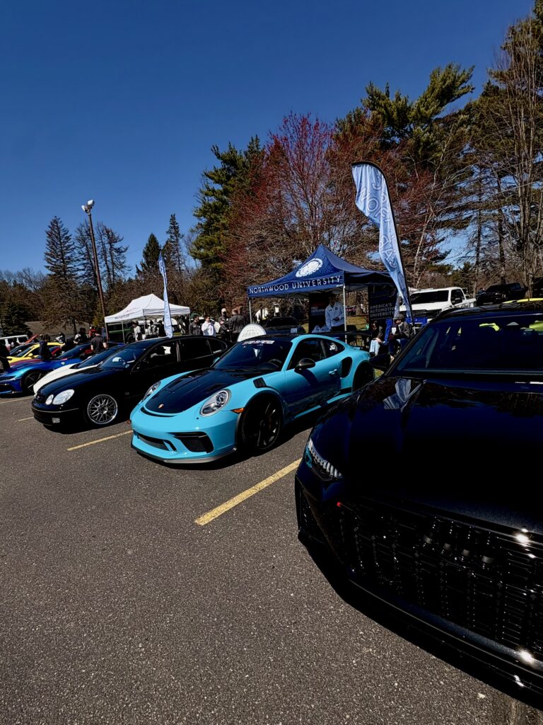 A row of sports cars, including a blue Porsche, are parked at an outdoor car show under clear skies with trees and tents in the background.