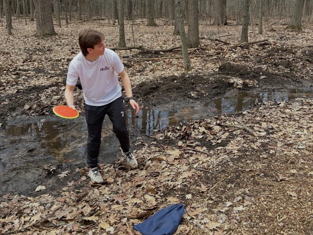 A person stands on leafy ground by a muddy stream in a forest, holding a disc in one hand and looking to the left.