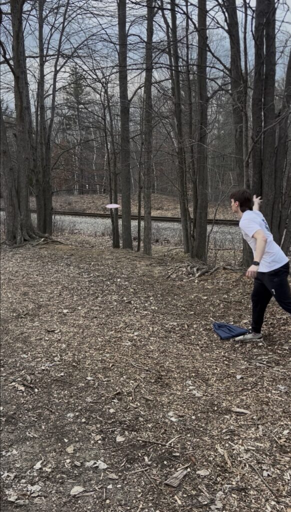 A person throws a disc through a wooded area on a disc golf course, with trees and a railroad track in the background.