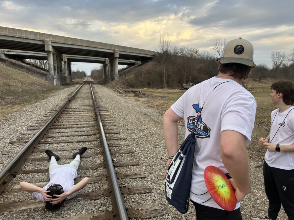 Three young people are next to train tracks, with one lying on the tracks and two standing nearby. A bridge crosses over the tracks in the background.