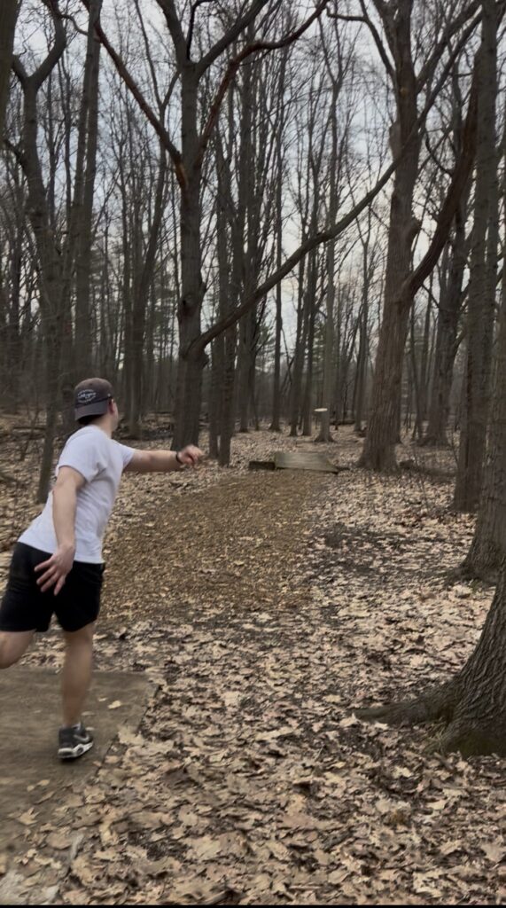 A person in a white shirt and black shorts throws a disc on a wooded disc golf course covered with fallen leaves.