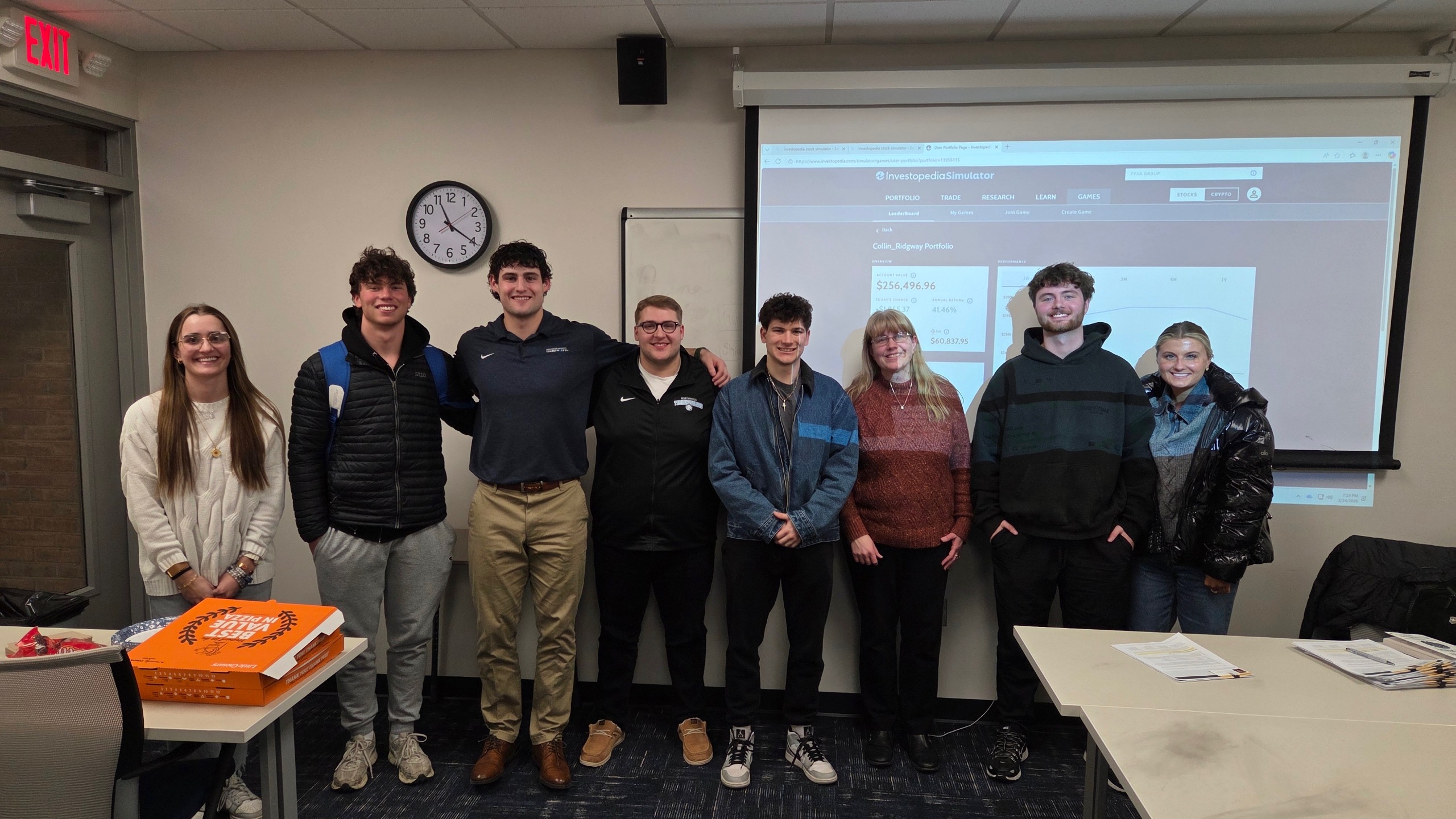 Eight people stand in a row, smiling for a group photo in a classroom at the FFAA meeting. A table with pizza boxes and a projector screen are visible in the background.