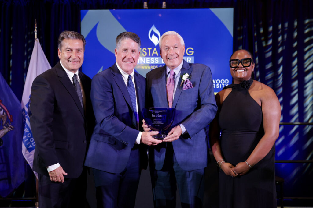 Four people in formal attire stand on stage; two men in the center hold a blue glass award, while two others, a man and a woman, stand on either side of them.