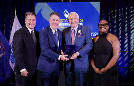 Four people in formal attire stand on stage; two men in the center hold a blue glass award, while two others, a man and a woman, stand on either side of them.