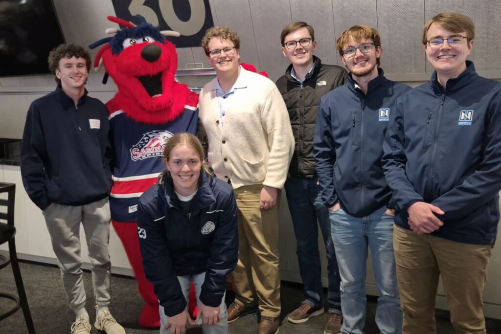 A group of six young adults from the Honorary Accounting Society of Northwood University pose indoors with a person in a red mascot costume wearing a hockey jersey.