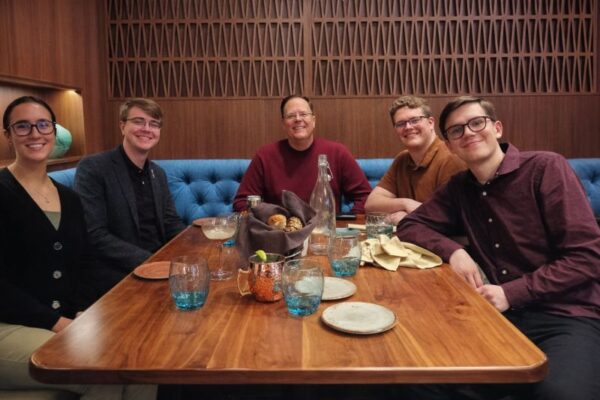 Five people from the Honorary Accounting Society of Northwood University are seated around a wooden dining table in a restaurant, smiling at the camera with plates, glasses, and a bread basket on the table.