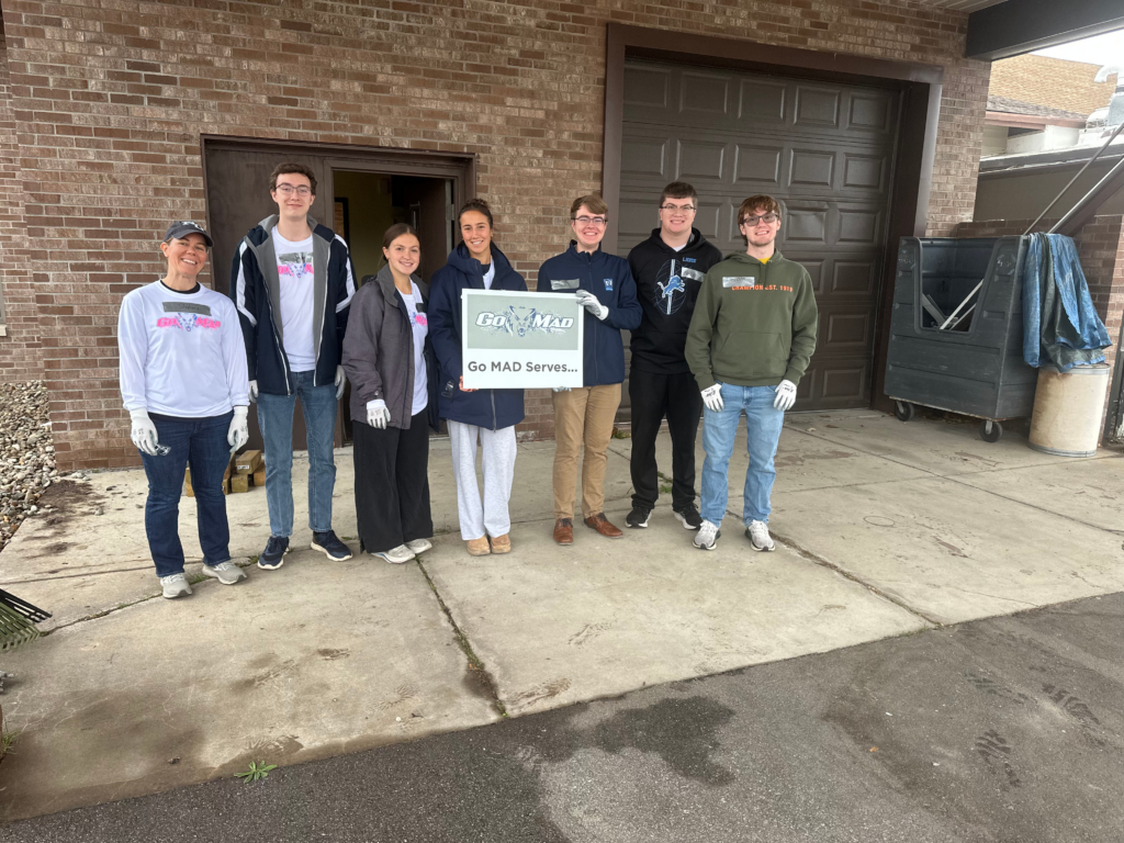 Seven people stand outside a brick building, some wearing gloves, holding a "Go MAD Serves" sign. Gardening tools and a dumpster are visible nearby, as members of the Honorary Accounting Society of Northwood University volunteer together.