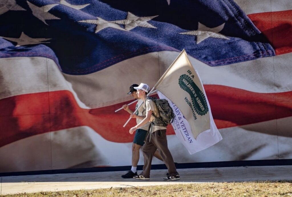 Two people walk in front of a large American flag mural, one carrying a flag that reads “Military & Veteran Powered,” “Pushups for Patriots,” and features the Kappa Sigma logo.