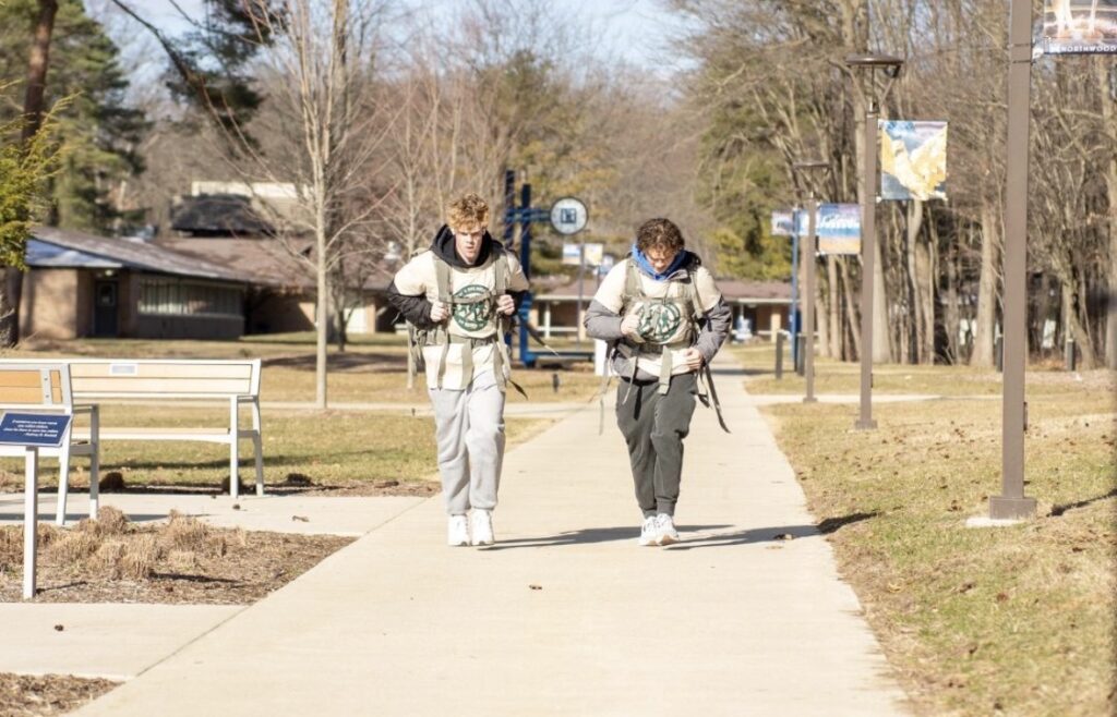 Two people wearing backpacks and weighted vests, possibly members of Kappa Sigma, walk side by side on a paved campus pathway on a sunny day.