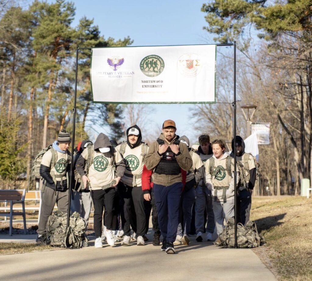A group of people in matching shirts and backpacks, some displaying Kappa Sigma pride, walk under a banner for Northwood University on a sunny outdoor path, surrounded by trees.