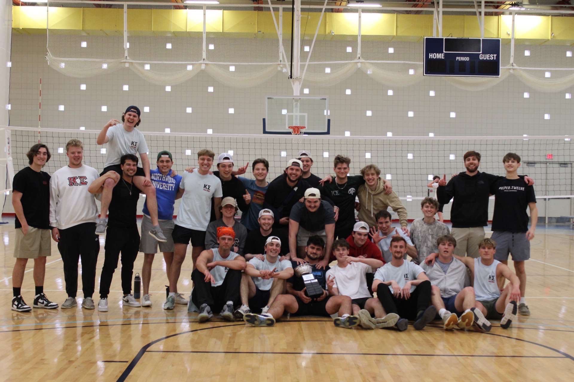 A large group of young Kappa Sigma men pose together on an indoor basketball court in front of a volleyball net, some standing and some sitting, proudly holding a trophy.