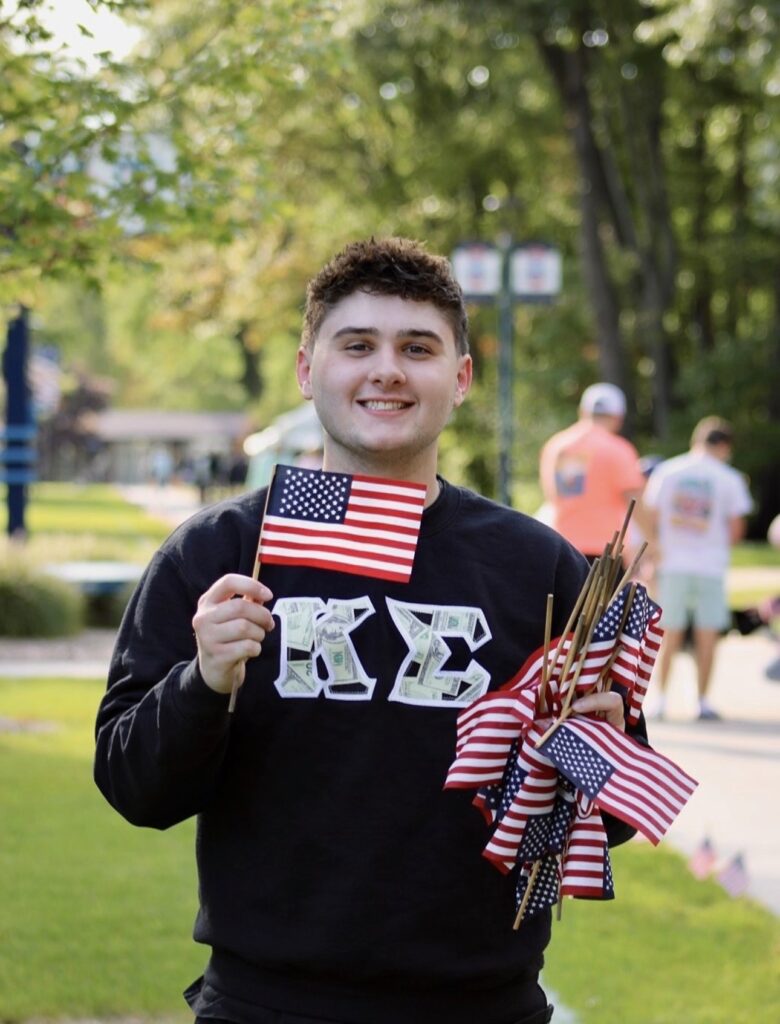 A young man holding several small American flags and wearing a black Kappa Sigma sweatshirt with Greek letters stands outside on a sunny day.
