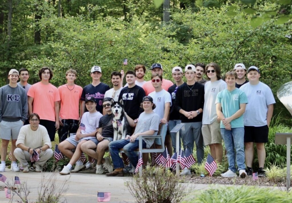 A group of young men and a dog, representing Kappa Sigma, pose outdoors with American flags placed on the ground around them. Trees and greenery are in the background.