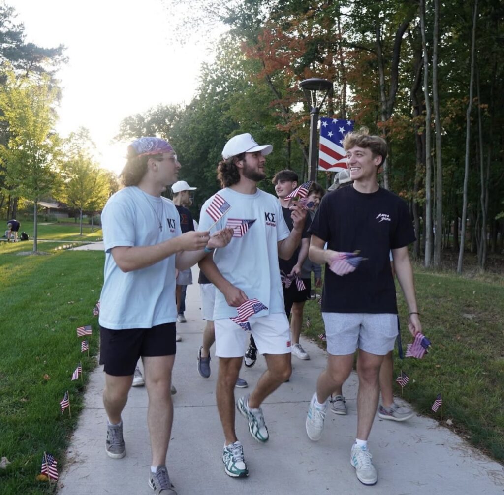 A group of young Kappa Sigma men walk on a sidewalk lined with small American flags, holding flags and talking in a park setting.