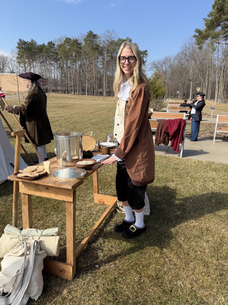 Isabella Stephens, dressed in colonial-style clothing, stands at a wooden table with bread, bowls, and a metal pot outdoors near Washington D.C., as others gather nearby among the trees.