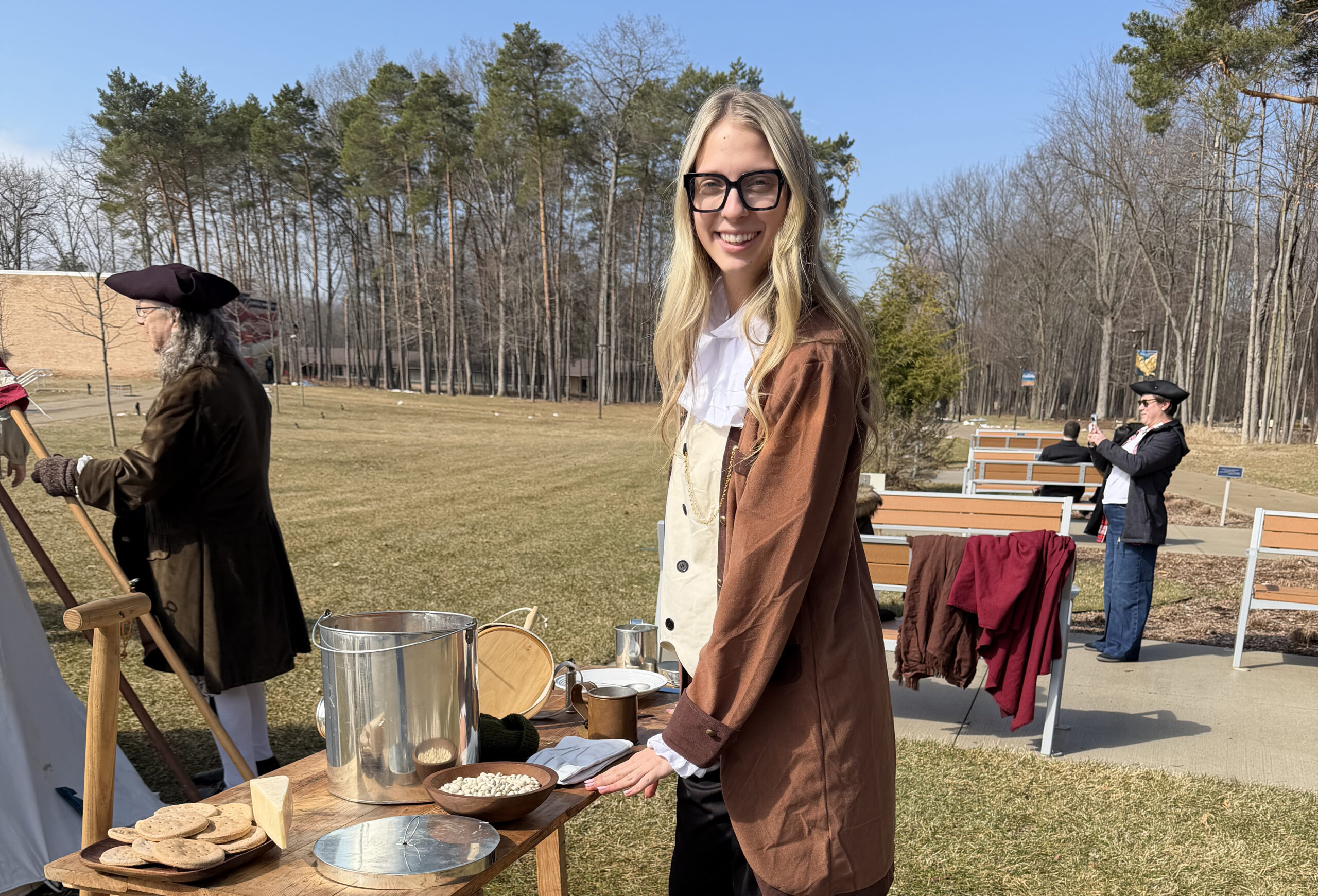 Isabella Stephens, dressed in historical attire, stands at an outdoor table with food items, while two others in colonial costumes are in the background. Trees and benches fill the Washington D.C. scene during Translate the Declaration.