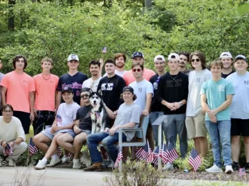 A group of young men from student organizations and a dog pose outdoors, some sitting and some standing, with small American flags and greenery in the background.