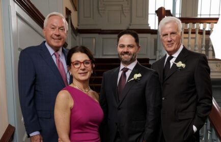 Four formally dressed adults, including three men in suits with boutonnieres and a woman in a sleeveless magenta dress, stand together indoors before a staircase—distinguished as Northwood University Business Leaders from the class of 2026.