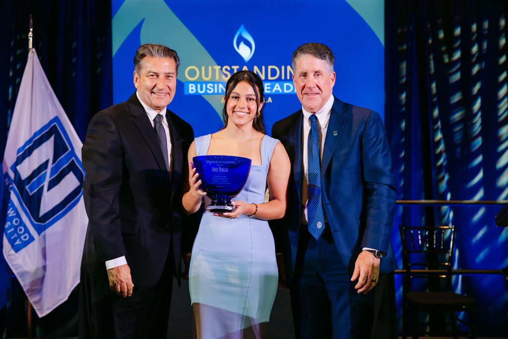 Three people in formal attire stand on stage; the woman in the center holds an award. A sign behind them reads "Outstanding Business Lead.