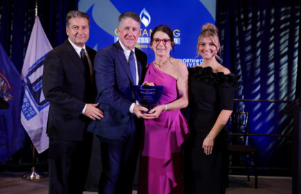 Four people in formal attire stand together on stage, holding an award, with flags and a presentation screen in the background.