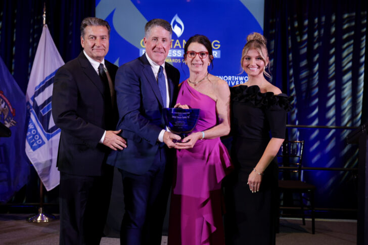 Four people in formal attire stand together on stage, holding an award, with flags and a presentation screen in the background.