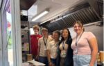 Five young people stand together inside a food truck, posing for a photo. They are wearing casual clothes and lanyards, with kitchen equipment visible behind them.