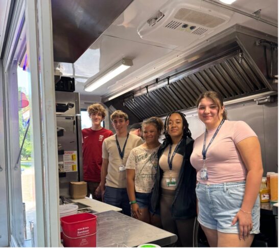 Five young people stand together inside a food truck, posing for a photo. They are wearing casual clothes and lanyards, with kitchen equipment visible behind them.