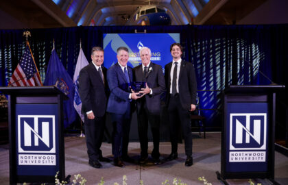 Four men in suits stand on a stage at Northwood University, one holding an award plaque, with flags, podiums, and a blue backdrop behind them.