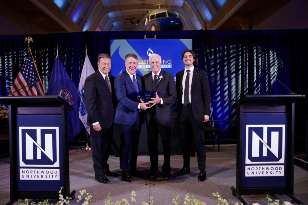Four men in suits stand on stage at the 2026 Northwood University Outstanding Business Leaders event, holding an award, with flags and a jet in the background.