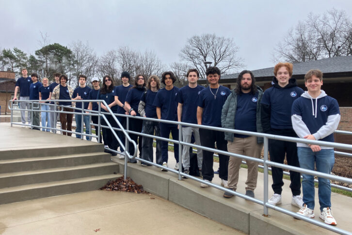 A group of students wearing matching navy blue t-shirts stands in a line along a ramp outdoors on a cloudy day, representing Northwood University’s commitment to Cybersecurity excellence.