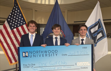 Three young men in suits hold a large $10,000 check from Northwood University for the Disc Golf Club, standing in front of American and university flags.