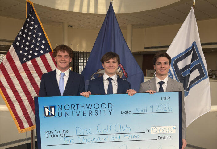 Three young men in suits hold a large $10,000 check from Northwood University for the Disc Golf Club, standing in front of American and university flags.