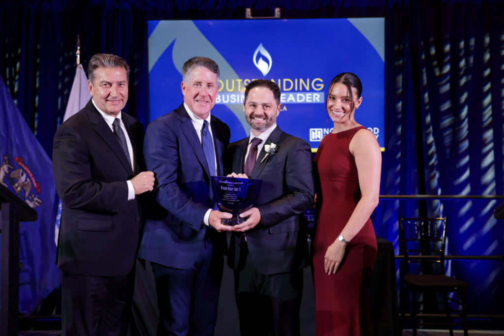 Four people in formal attire stand on stage; two men in the center hold an award, with a screen behind them reading "Outstanding Business Leader.