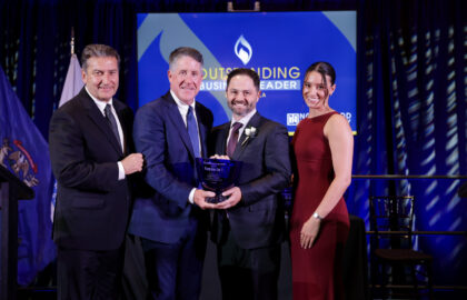Four people in formal attire stand on stage; two men in the center hold an award, with a screen behind them reading "Outstanding Business Leader.