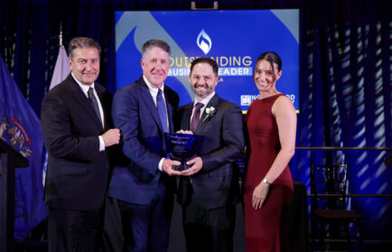 Four people in formal attire stand on stage; two men in the center hold an award, with a screen behind them reading "Outstanding Business Leader.