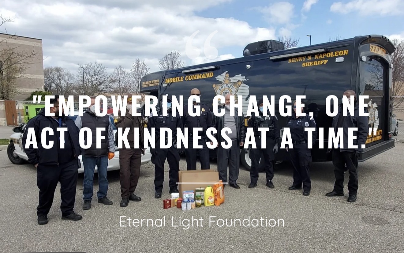 A group of uniformed officers stands by a police vehicle with boxes of food on the ground; large text reads, "EMPOWERING CHANGE, ONE ACT OF KINDNESS AT A TIME," shining as an Eternal Light in the community.