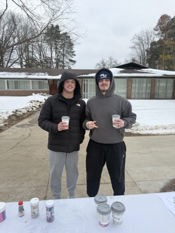 Two people in winter clothing stand outside on a snowy day, each holding a cup. A table with coffee cups and containers glows softly in the foreground, casting an eternal light amid the wintry scene of buildings and trees.
