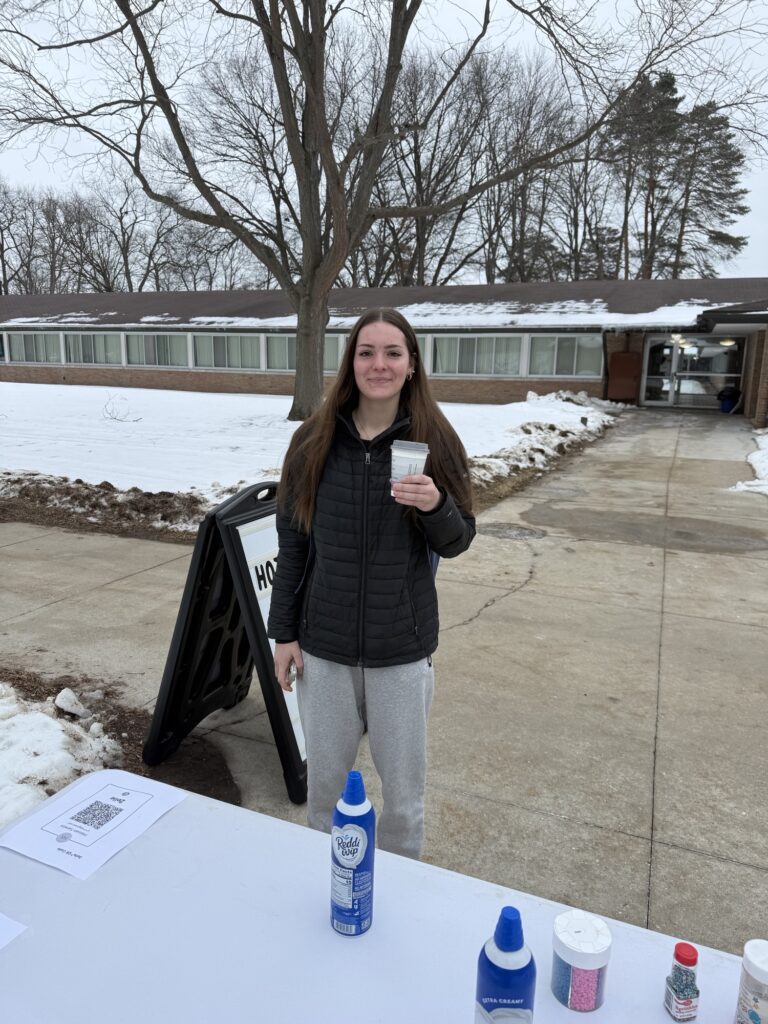 A young woman in a black jacket and gray sweatpants holds a cup outside on a snowy sidewalk near a table with whipped cream, sprinkles, and an "Eternal Light" sign.