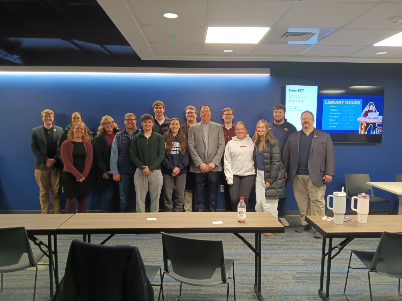 A group of people stands together in a conference room, posing for a photo in front of a blue wall with a screen displaying library hours, representing the Honorary Accounting Society of Northwood University.